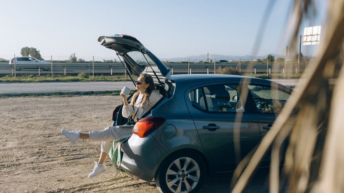 A woman sitting in the trunk of a parked car on a sunny day, enjoying a break during a road trip.